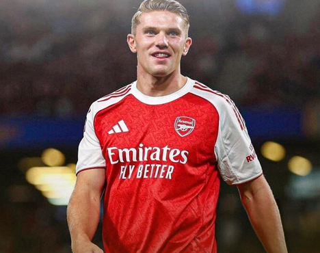 Спортсмен в футбольной форме команды Арсенал стоит на поле. Красная футболка с белыми рукавами, логотип клуба и спонсорская надпись видны на форме. Фон размыт, с огнями стадиона